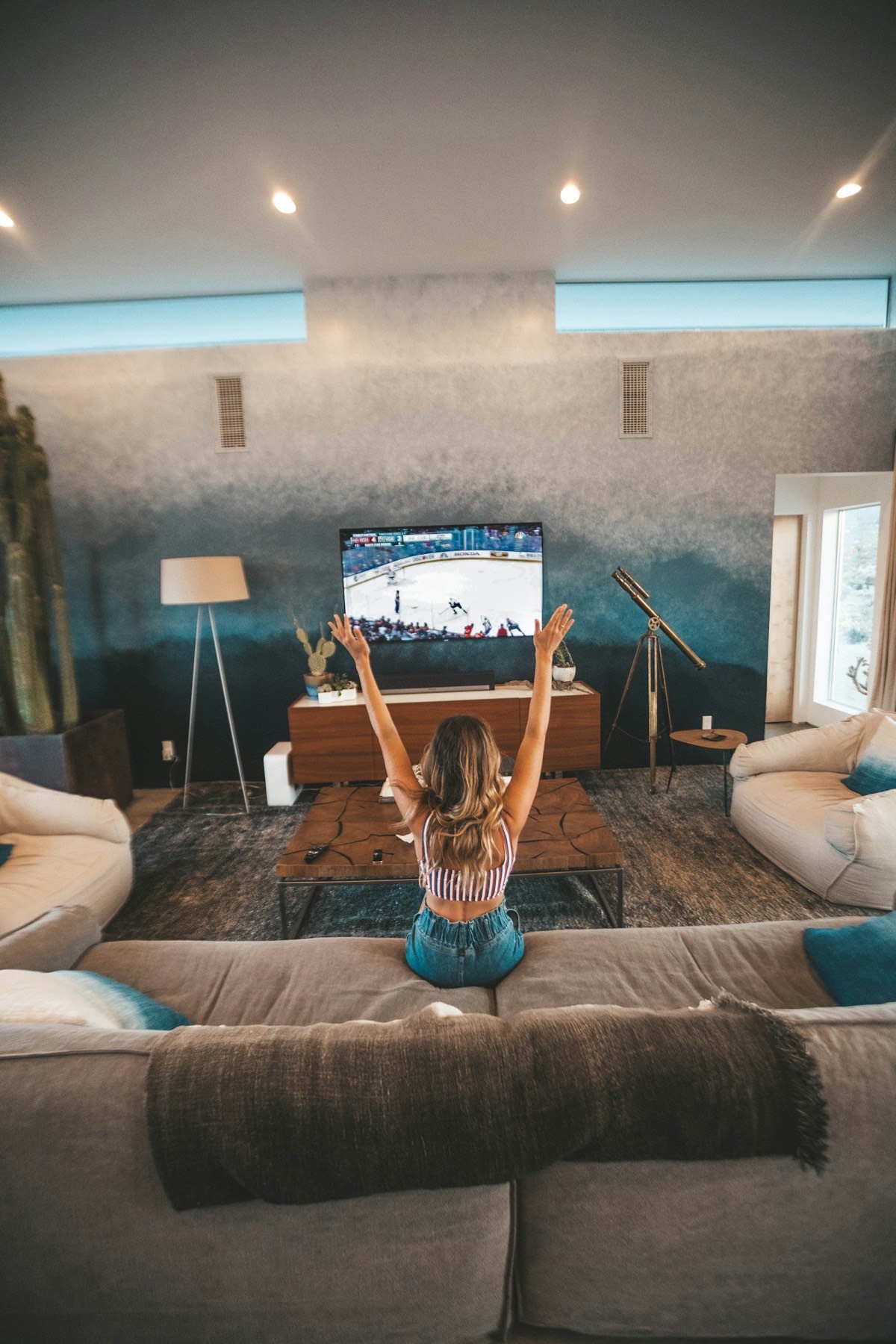 Woman celebrating with raised arms while watching a live sports broadcast on a wall-mounted television at home