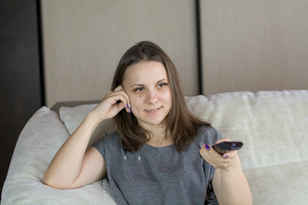 Woman relaxing on a couch at home, holding a TV remote control while choosing what to watch